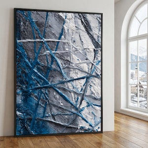 Puede incluir: Pintura abstracta texturizada en un marco negro. La obra presenta una mezcla de tonos blancos, grises y azules, con pinceladas visibles que crean un efecto dinámico y en capas. La pintura se exhibe en una pared con suelo de madera.