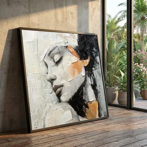 May include: A framed abstract painting of a woman's face in profile. The artwork uses a palette of white, gray, black, and orange. The woman's eyes are closed, and the style is textured, with visible brushstrokes. The frame is black.