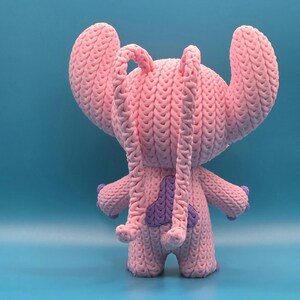 May include: A handmade pink knitted plush toy with large ears and a purple accent on the back. The toy has a textured appearance and is standing on a blue surface. The toy is a character from a popular animated film.