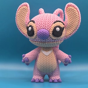 May include: A pink and purple figurine of the character Angel from the Lilo & Stitch franchise. The figurine has large ears, black eyes, and a white heart-shaped detail on its chest. The figure is made of a textured material, giving it a knitted appearance.