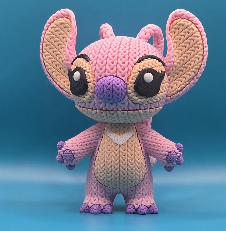 May include: A pink and purple figurine of the character Angel from the Lilo & Stitch franchise. The figurine has large ears, black eyes, and a white heart-shaped detail on its chest. The figure is made of a textured material, giving it a knitted appearance.