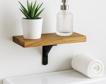 May include: A wooden shelf with a black metal bracket, holding a small potted plant, a clear glass soap dispenser, and a rolled white towel. The shelf is mounted on a white wall above a white toilet.