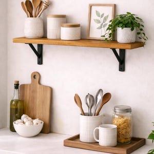 May include: A kitchen scene with a wooden shelf holding ceramic jars, wooden spoons, and a framed botanical print. Below, a cutting board, bowl of garlic, and a wooden tray with utensils and a mug. A jar of pasta is also visible.