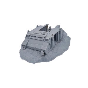 May include: A gray miniature tank model with detailed features, including tracks, hatches, and a sloped front. The model is set on a textured, gray base, suggesting a terrain or diorama setting. The overall design is reminiscent of a military vehicle.