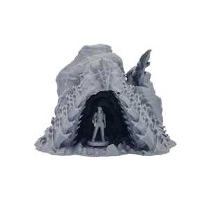 Puede incluir: Una cueva en miniatura gris impresa en 3D con una figura en su interior. La cueva tiene un exterior dentado y texturizado y un interior oscuro. La figura es una pequeña representación detallada de una persona.