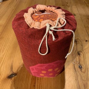 Peut inclure: Un sac cylindrique en tissu rouge avec une fermeture à cordon. Le sac présente un motif patchwork avec des accents orange et rose. Le cordon de serrage est blanc et la doublure intérieure est d'une teinte orange plus claire. Le sac est posé sur une surface en bois.