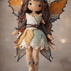 May include: A handmade crochet fairy doll with brown hair, large eyes, and a floral headpiece. The doll wears a layered dress in white, yellow, and blue, with matching wings. The doll's legs are tan with colorful leaf accents.