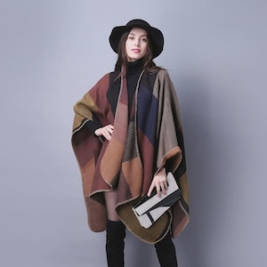 May include: A woman wearing a multi-colored poncho with a geometric pattern in shades of brown, red, blue, and olive green. She is also wearing a black hat, black turtleneck, black boots, and carrying a black and white clutch.