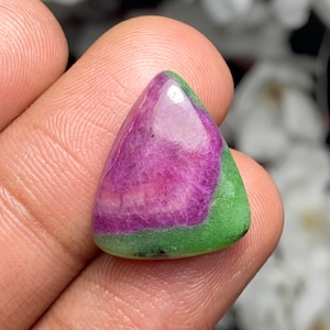 Puede incluir: Una gema triangular pulida con una vibrante paleta de colores. La piedra presenta una banda central de magenta intenso, bordeada por un borde verde. La gema se sostiene en una mano, mostrando su superficie lisa y reflectante.