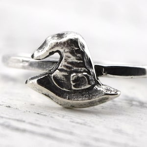 May include: A silver-toned metal ring featuring a small, three-dimensional witch hat charm centered on the band. The metal has a darkened, oxidized finish in the recessed areas of the hat design, highlighting the pointed top and wide brim.