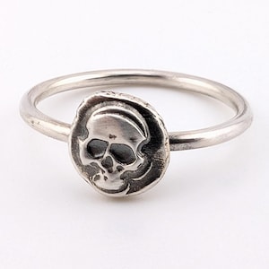May include: A silver ring featuring a skull design. The skull is centered on a circular, textured setting. The ring band is a simple, polished silver. The skull has detailed eye sockets, nose, and teeth.