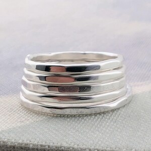 May include: A stack of five individual silver rings, each featuring a smooth, polished finish with a slightly irregular, organic shape. The bands are layered vertically, creating a cohesive set of metallic, reflective circular jewelry pieces.