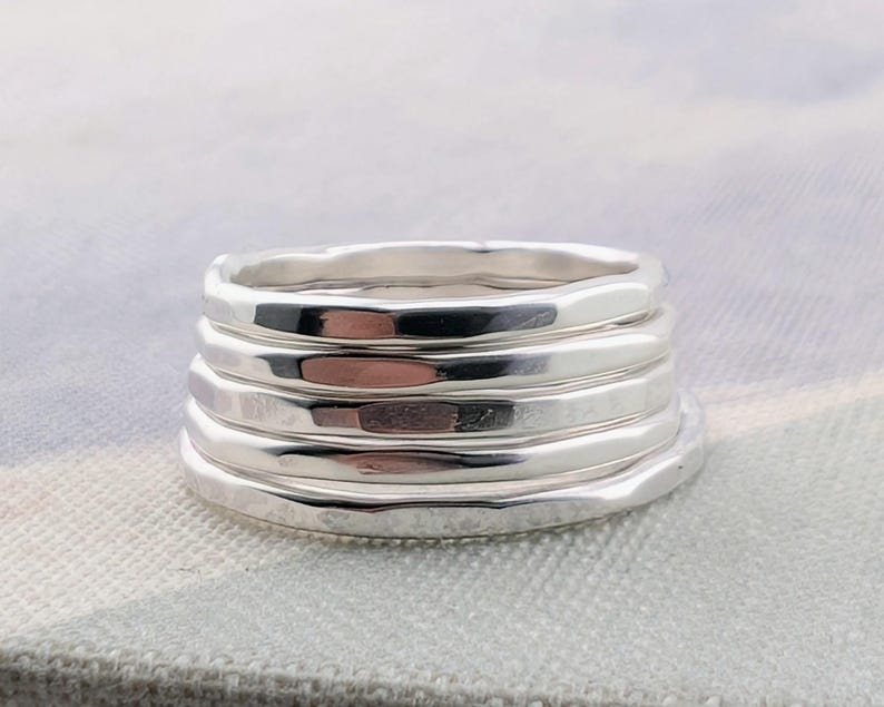 May include: A stack of five individual silver rings, each featuring a smooth, polished finish with a slightly irregular, organic shape. The bands are layered vertically, creating a cohesive set of metallic, reflective circular jewelry pieces.