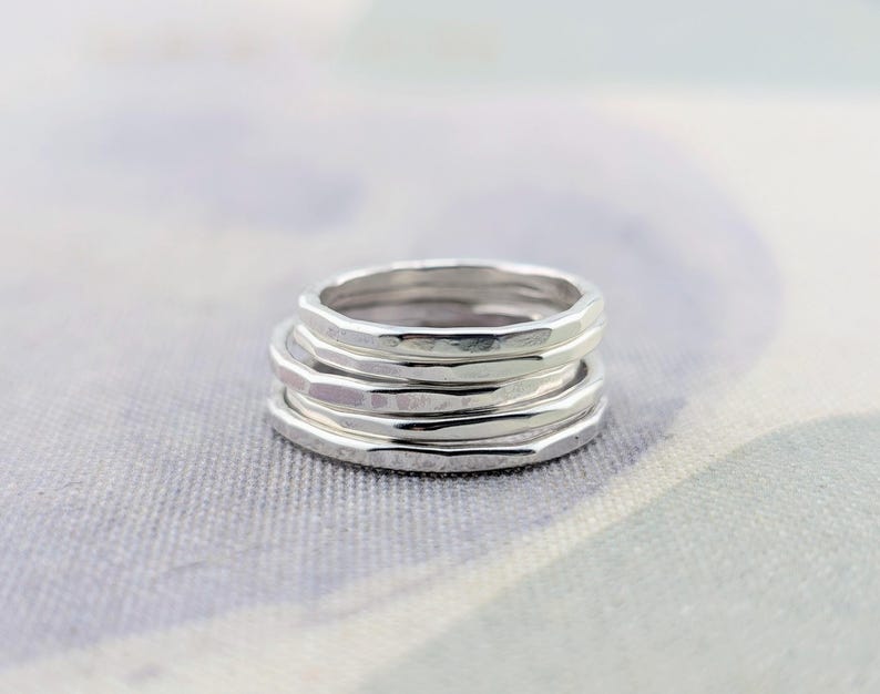 May include: A stack of three silver rings, each featuring a distinct hammered texture that creates a series of small, reflective facets along the outer surface of the metal bands. The rings are positioned one on top of the other in a vertical column.