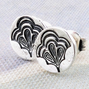 May include: A pair of circular silver stud earrings featuring a stamped, fan-shaped botanical motif with oxidized black detailing in the recessed areas. The metal surface has a slightly textured, matte finish with a post back closure mechanism.