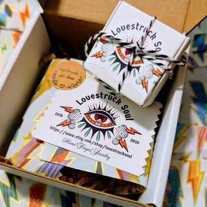 May include: A cardboard shipping box containing a small white jewelry box tied with black and white twine, a rectangular card with the text Lovestruck Soul, an eye graphic, and lightning bolts, and a round sticker reading handmade with love.