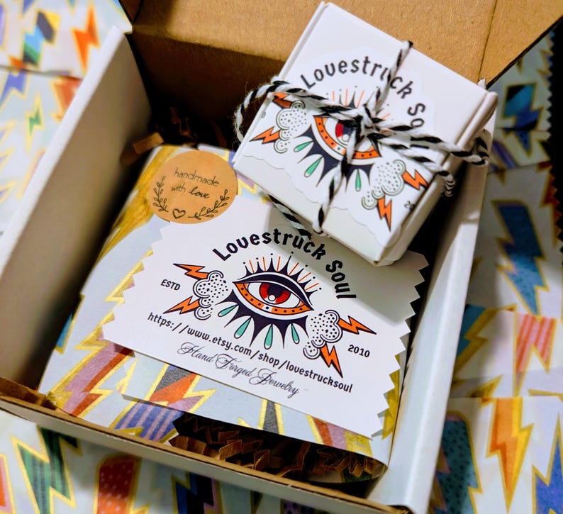 May include: A cardboard shipping box containing a small white jewelry box tied with black and white twine, a rectangular card with the text Lovestruck Soul, an eye graphic, and lightning bolts, and a round sticker reading handmade with love.