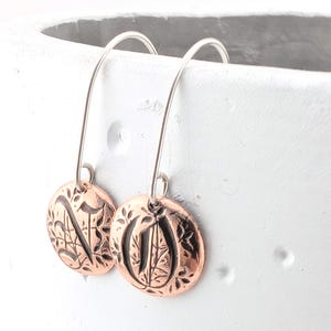 May include: A pair of drop earrings featuring circular copper-toned charms with an embossed botanical leaf pattern and dark recessed details. The charms are suspended from thin, curved silver-toned metal ear wires that hook through the top.