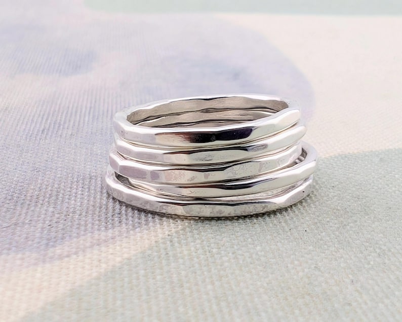 May include: A stack of five silver-colored rings with a hammered, textured finish. Each ring features a slightly irregular, organic shape with a polished, reflective surface. The rings are stacked neatly on top of one another in a vertical column.