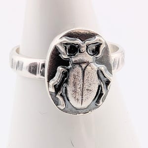 May include: A silver ring featuring an oval-shaped face with a detailed insect design. The insect has glasses and is centered on the ring's face. The band of the ring has a textured design.