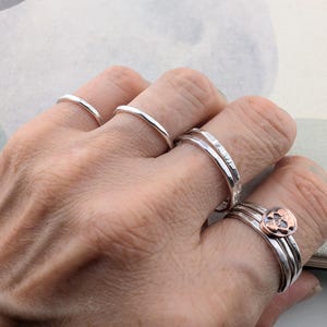 May include: A collection of silver rings worn on four fingers. The rings feature various textures, including smooth, hammered, and faceted bands. One ring on the ring finger is topped with a small, circular copper-toned skull emblem.