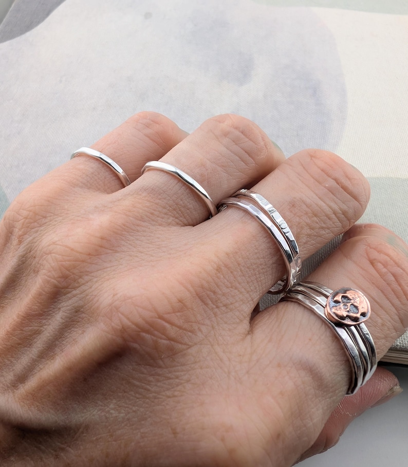 May include: A collection of silver rings worn on four fingers. The rings feature various textures, including smooth, hammered, and faceted bands. One ring on the ring finger is topped with a small, circular copper-toned skull emblem.