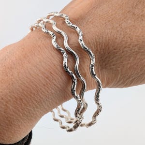 Puede incluir: Tres brazaletes plateados con un diseño ondulado y texturizado se muestran en una muñeca. Los brazaletes tienen una textura martillada y una forma orgánica y fluida. La joyería está hecha de un material metálico brillante.