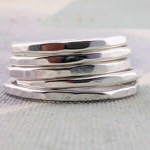 May include: A stack of five individual silver rings, each featuring a distinct hammered texture on the outer surface. The rings are polished to a high shine, creating a reflective metallic finish that catches the light across the uneven, dimpled metal bands.