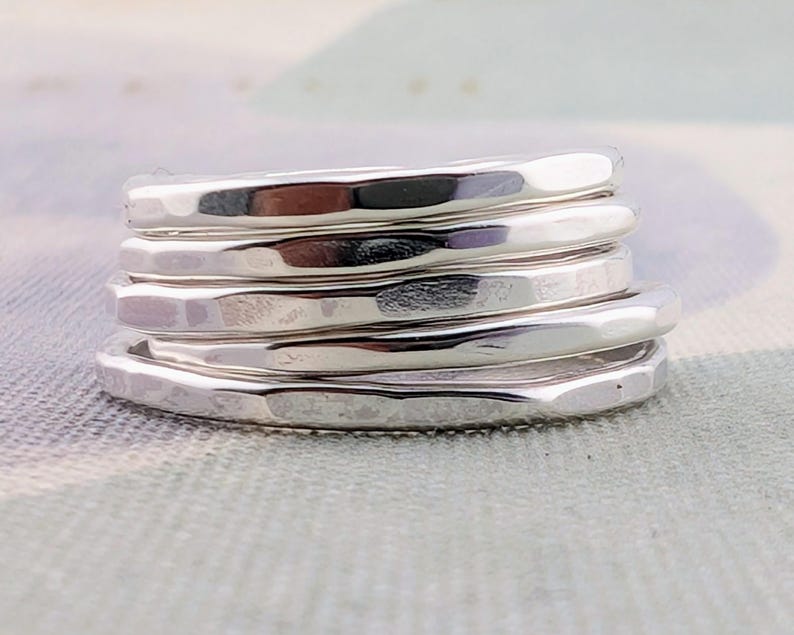 May include: A stack of five individual silver rings, each featuring a distinct hammered texture on the outer surface. The rings are polished to a high shine, creating a reflective metallic finish that catches the light across the uneven, dimpled metal bands.
