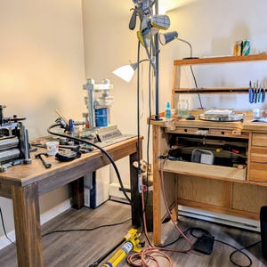 May include: A jewelry workbench setup featuring a wooden table with a metal rolling mill, a hydraulic press, and a second wooden jeweler's bench with drawers, hanging pliers, and various tools. A tall floor lamp stands between the two workstations.