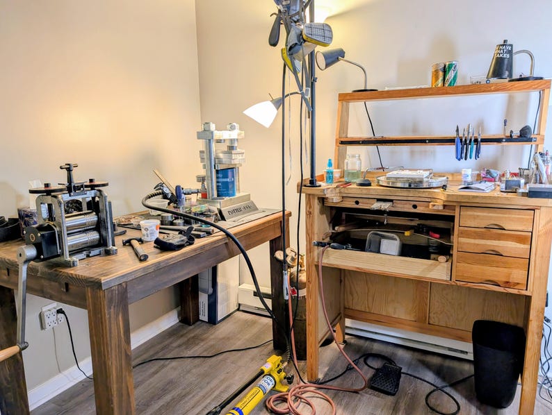 May include: A jewelry workbench setup featuring a wooden table with a metal rolling mill, a hydraulic press, and a second wooden jeweler's bench with drawers, hanging pliers, and various tools. A tall floor lamp stands between the two workstations.