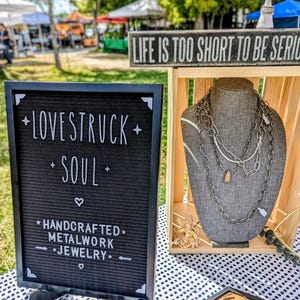 May include: A black felt letter board with white text reading LOVESTRUCK SOUL HANDCRAFTED METALWORK JEWELRY next to a wooden display stand holding a gray fabric bust wearing multiple layered silver-toned metal chain necklaces with a teardrop pendant.