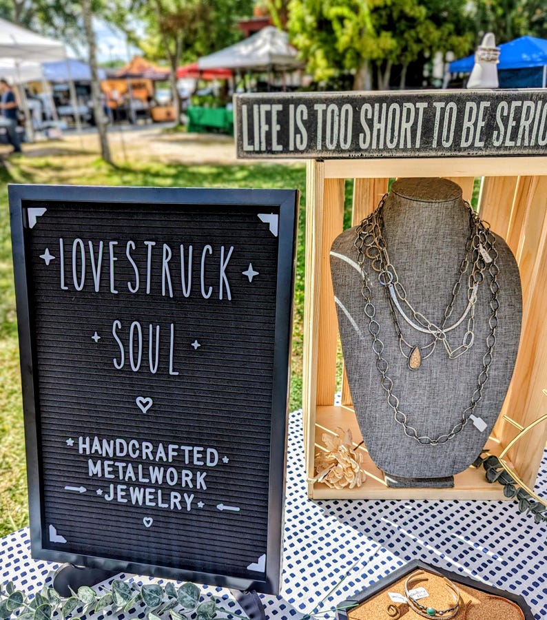 May include: A black felt letter board with white text reading LOVESTRUCK SOUL HANDCRAFTED METALWORK JEWELRY next to a wooden display stand holding a gray fabric bust wearing multiple layered silver-toned metal chain necklaces with a teardrop pendant.