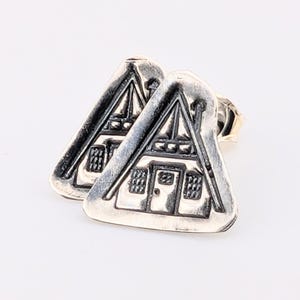 May include: A pair of silver-toned stud earrings. Each earring is shaped like a small house with a triangular roof and detailed windows and a door. The earrings are set against a white background.