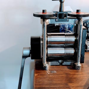May include: A manual metal rolling mill featuring a textured grey frame, two polished steel rollers with grooved sections, a black side-mounted crank handle, and a top-mounted adjustment T-handle with two black gear wheels with red accents.