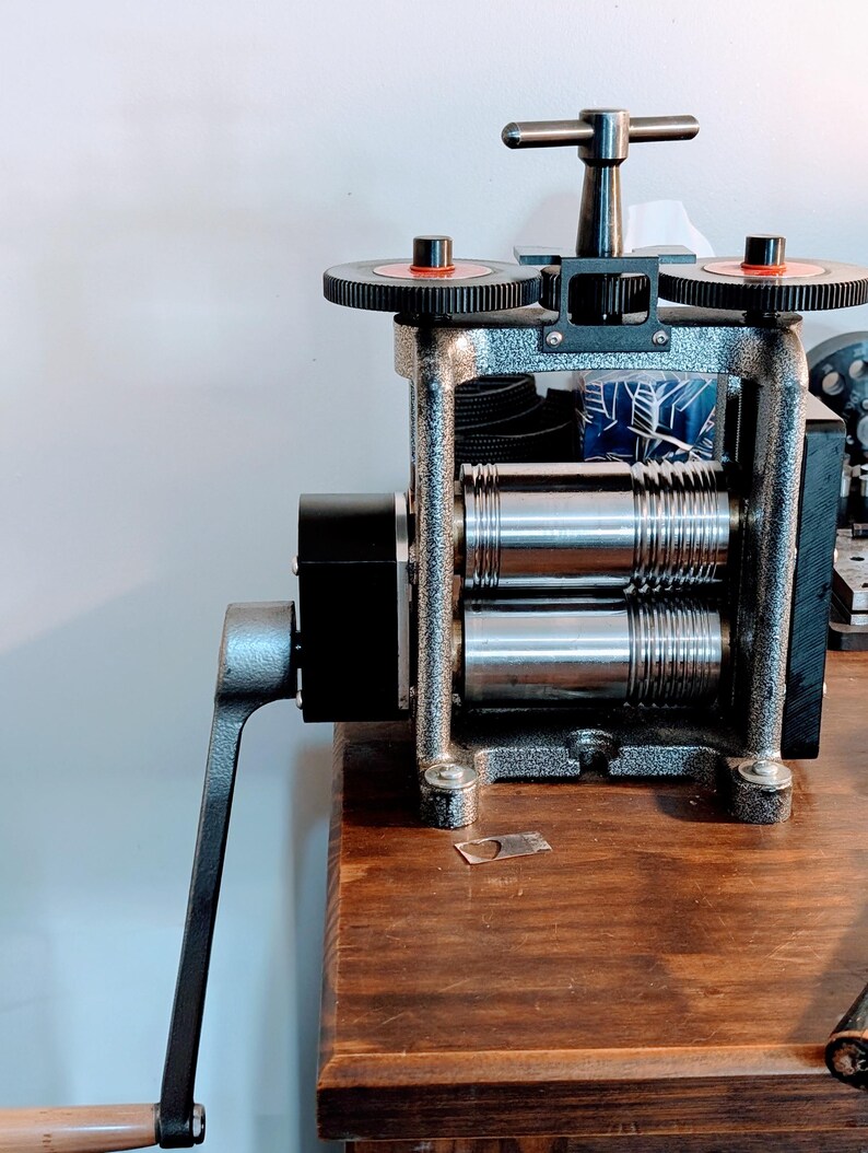 May include: A manual metal rolling mill featuring a textured grey frame, two polished steel rollers with grooved sections, a black side-mounted crank handle, and a top-mounted adjustment T-handle with two black gear wheels with red accents.