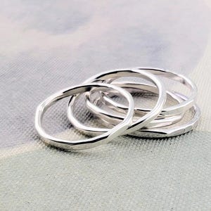 May include: A set of four silver-colored rings with a smooth, polished finish and a slightly irregular, organic circular shape. The rings are stacked together, showcasing their reflective metallic surface and simple, continuous band design.