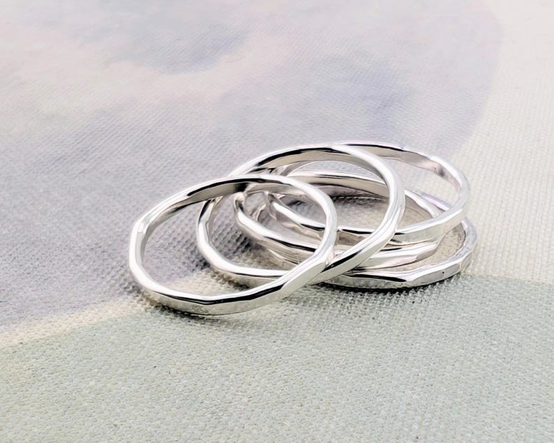 May include: A set of four silver-colored rings with a smooth, polished finish and a slightly irregular, organic circular shape. The rings are stacked together, showcasing their reflective metallic surface and simple, continuous band design.
