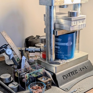 May include: A Potter USA hydraulic press featuring a blue Temco Industrial hydraulic cylinder, a metal frame with two top nuts, and a base plate. The setup includes a digital caliper, a wooden ruler, and various metal tools in clear containers.