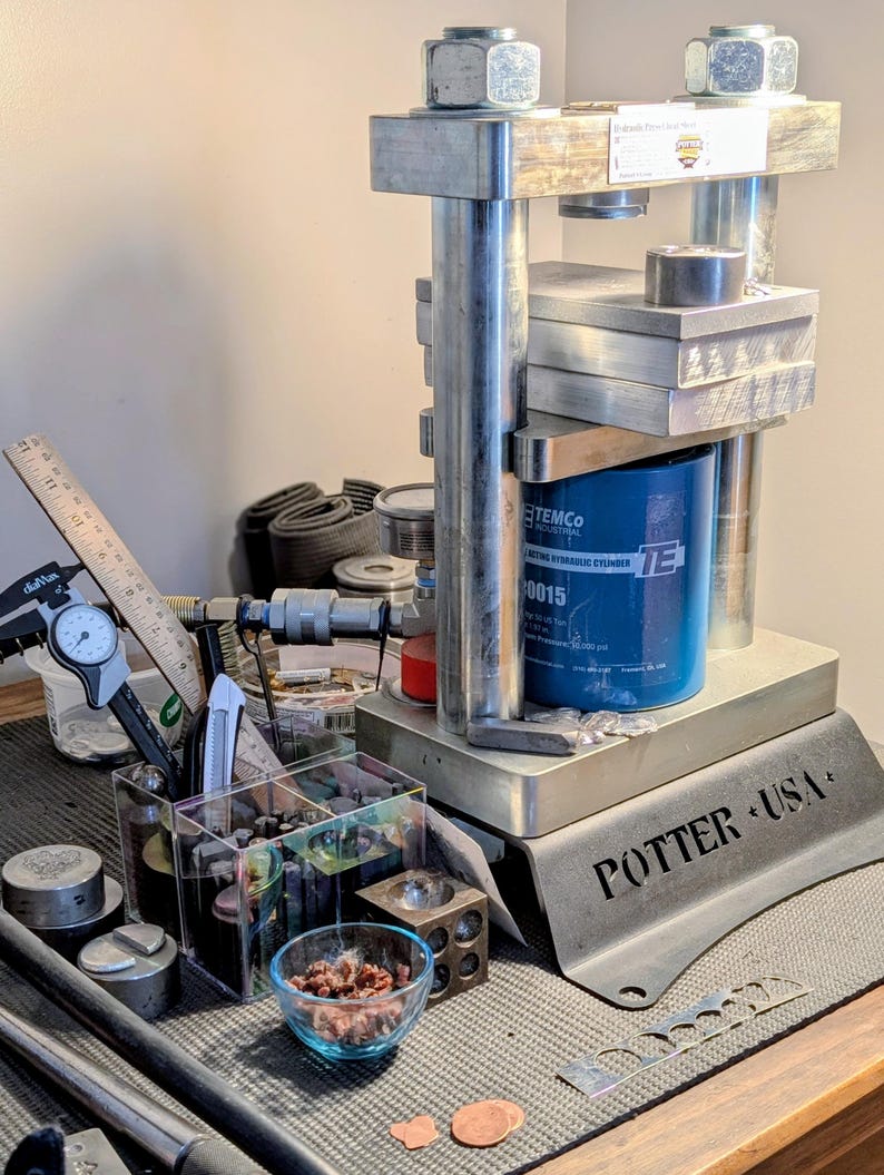 May include: A Potter USA hydraulic press featuring a blue Temco Industrial hydraulic cylinder, a metal frame with two top nuts, and a base plate. The setup includes a digital caliper, a wooden ruler, and various metal tools in clear containers.