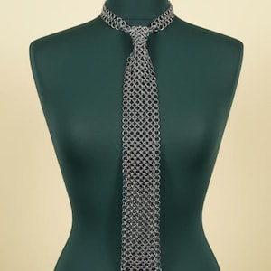 Puede incluir: Una corbata de cota de malla plateada exhibida en un maniquí verde. La corbata está hecha de pequeños anillos de metal entrelazados, creando una tela texturizada y flexible. La corbata está anudada en el cuello y cuelga, mostrando su intrincado diseño y artesanía.