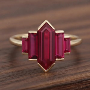 Puede incluir: Un anillo de oro con un rubí central hexagonal alargado, flanqueado por dos rubíes más pequeños de corte baguette a cada lado. Las cinco piedras preciosas de color rojo vibrante están engastadas en una banda de oro pulido, creando un diseño geométrico simétrico y escalonado.