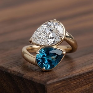 May include: A bypass ring crafted from polished yellow gold, featuring two pear-cut gemstones. One stone is a clear, colorless faceted gem, while the other is a deep blue faceted gemstone. The stones are set in a prong setting on a smooth band.