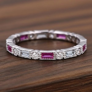 May include: A silver-toned eternity band featuring an alternating pattern of rectangular baguette-cut pink gemstones and round brilliant-cut clear stones. The metal setting displays a delicate milgrain texture along the edges of each stone.