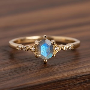 May include: A gold ring featuring a hexagonal, blue-grey gemstone at the center. The ring has small, clear stones on either side of the central stone and is set on a gold band. The ring is displayed on a wooden surface.