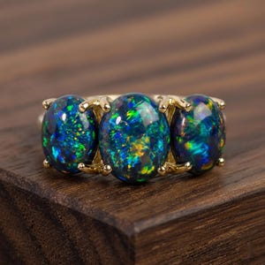 Könnte beinhalten: Ein Goldring mit drei ovalen, dunkelblauen Opalen in einer Reihe. Jeder Opal zeigt lebhafte grüne, gelbe und blaue Farbspiele innerhalb des Steins. Die Steine sind mit kleinen Goldkrallen auf einer polierten Goldschiene befestigt.