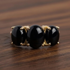 Può includere: Un anello dorato con tre grandi pietre nere ovali. Le pietre sono incastonate in una fascia dorata con griffe decorative. L'anello è esposto su una superficie di legno.