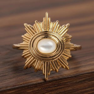 May include: A gold-colored ring featuring a sunburst design. The ring has a central oval-shaped, pearlescent stone. The sunburst design has pointed rays extending outwards from the center, creating a radiant effect. The ring is set against a wooden surface.