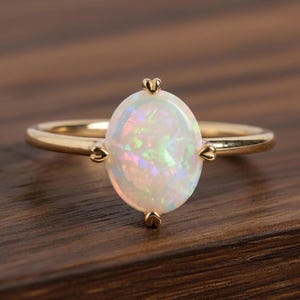 May include: A gold ring featuring an oval opal gemstone. The opal displays iridescent colors and is set with four heart-shaped prongs. The band is a simple, polished gold. The ring is displayed on a dark wood surface.