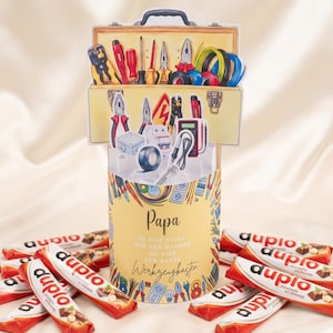 May include: A Father's Day gift set featuring a yellow toolbox-shaped card with illustrations of tools, a cylindrical card with the word "Papa" and text, and several chocolate bars with red and white packaging.